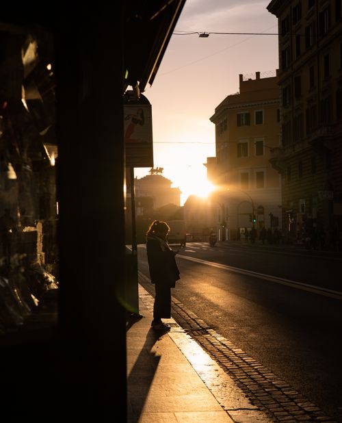 Free A woman stands silhouetted at sunset on a quiet city street, creating a serene urban scene. Stock Photo