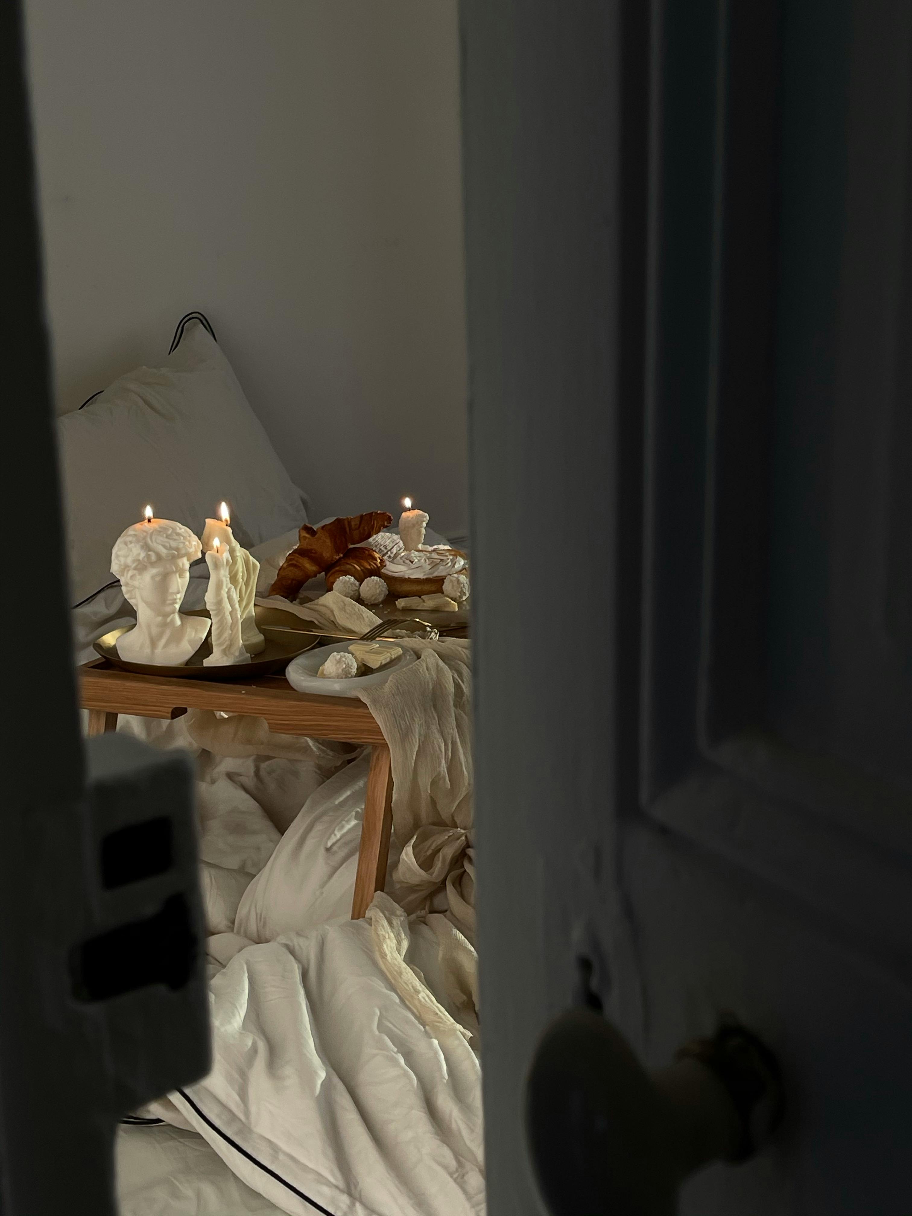 A tranquil bedroom scene with croissants, candles, and artistic sculptures on a wooden table.