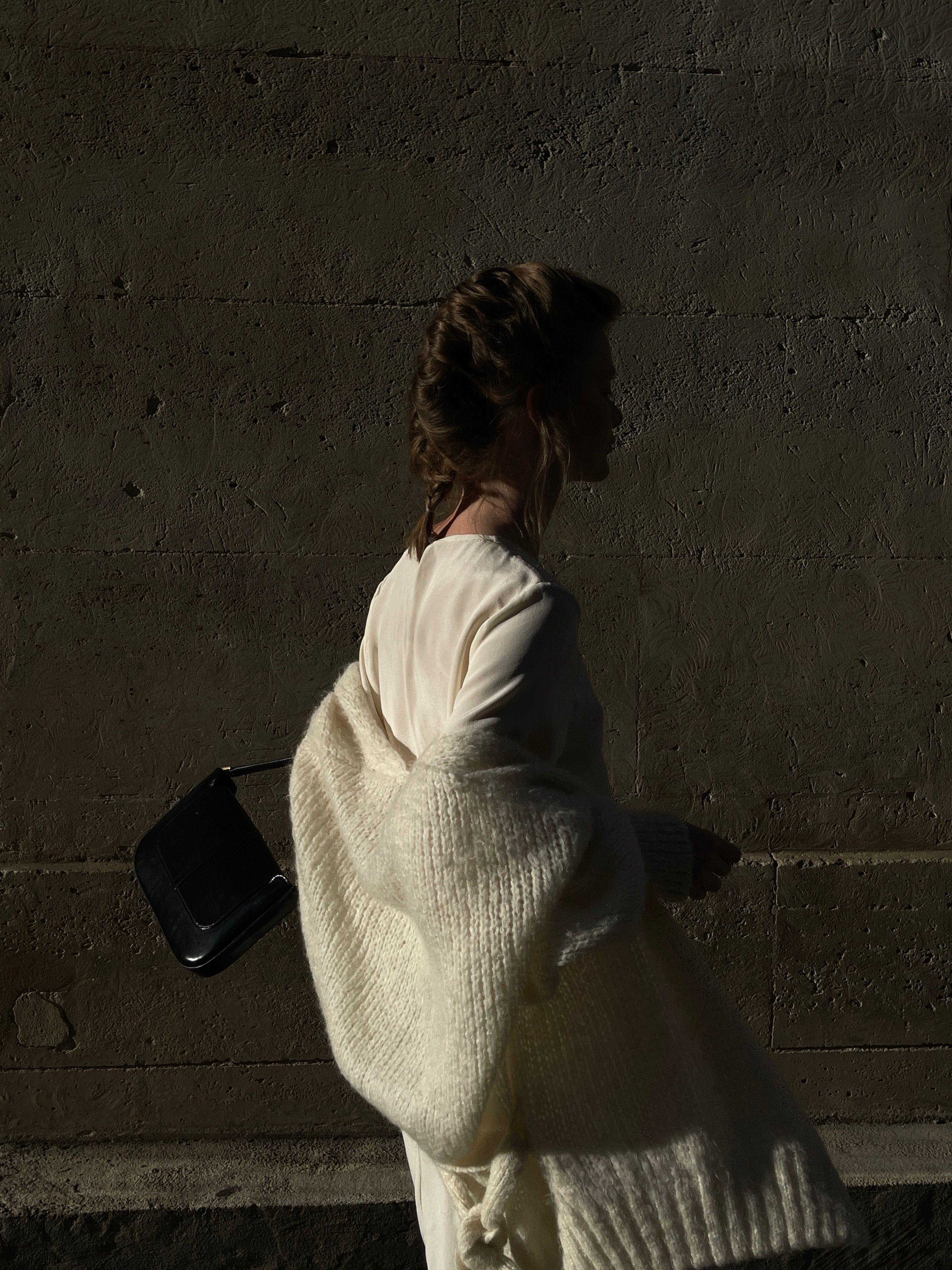 Stylish woman in white coat carrying handbag against urban wall.