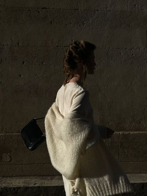 Stylish woman in white coat carrying handbag against urban wall.