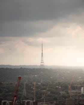 Aerial view of a television tower surrounded by cranes during dawn with clouded sky.