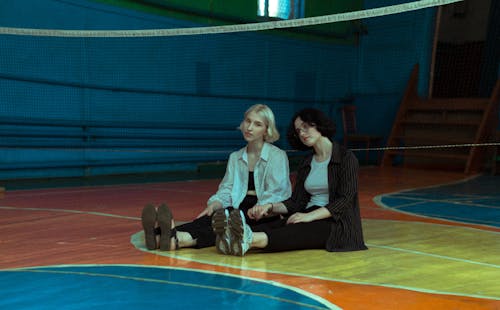 Two Young Women Sitting on a Floor in a School Gymnasium and Holding Hands 