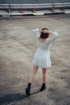 A woman in a white dress poses on an empty asphalt surface, surrounded by a moody urban setting.