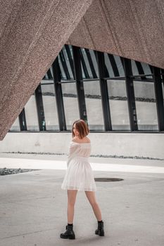 A young woman in a white dress poses stylishly under modern architectural structures outdoors.