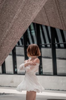 A young woman in a white dress posing gracefully in front of modern urban architecture.