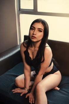 Sensual portrait of a woman in black lingerie sitting on a sofa by a window.