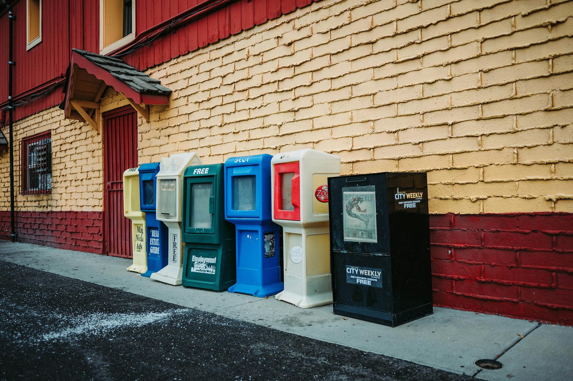 A variety of newspaper vending machines against a vibrant brick wall on the street.