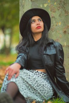 Stylish woman in a hat and black leather jacket posing in a park setting.
