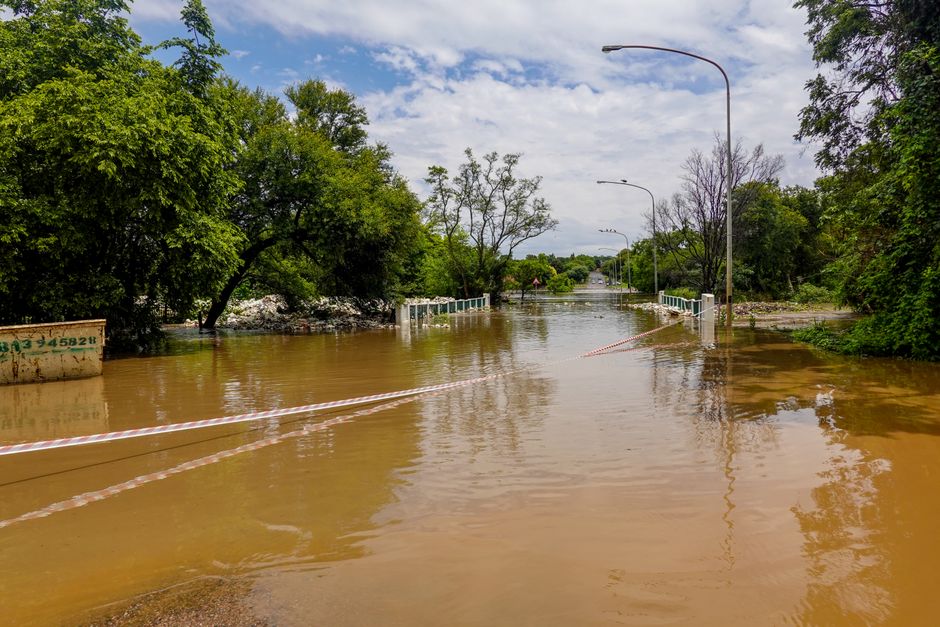Photo by Magda Ehlers A flooded road with a submerged bridge, surrounded by trees, under a cloudy sky.