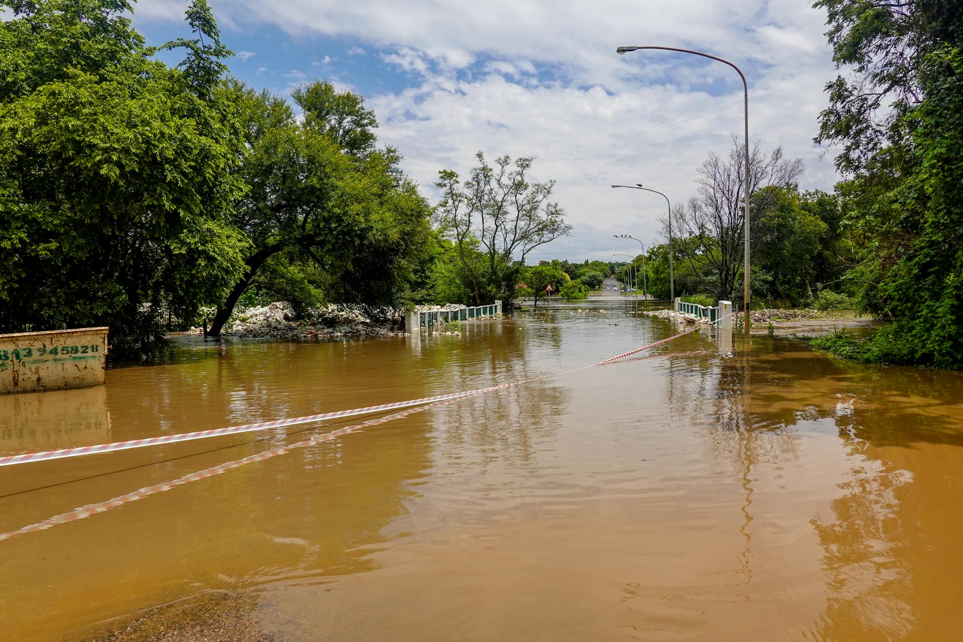 A flooded road with a submerged bridge, surrounded by trees, under a cloudy sky.