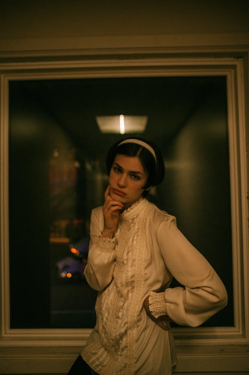 Woman in a Blouse and a Headband Standing by a Window with her Hands on her Hip and Face