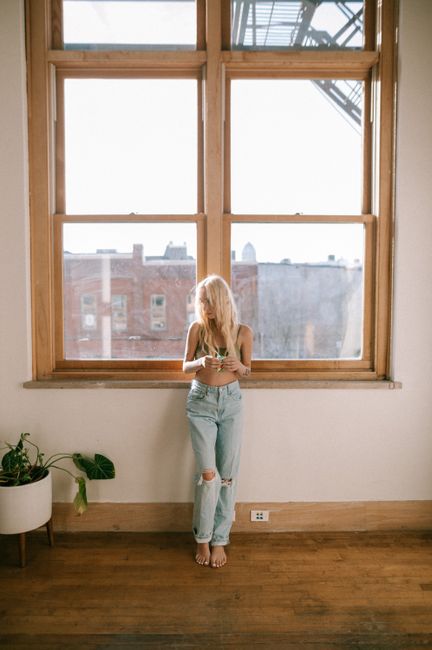 Blonde woman in casual jeans stands by a large indoor window, embracing natural light.