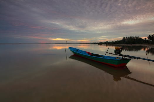 Tranquil sunset over a calm beach with a lone fishing boat casting reflections on the water.
