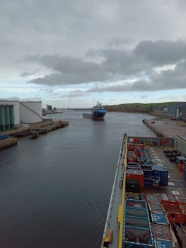A container ship approaches a bustling industrial port in England, showcasing logistics and maritime activity.