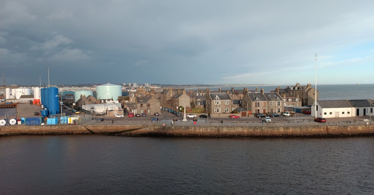 Overview of a coastal town's waterfront with buildings and industrial structures under a cloudy sky.