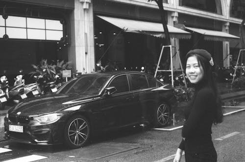 A Grayscale Photo of a Woman in Black Long Sleeves Standing Near the Car Parked on the Street