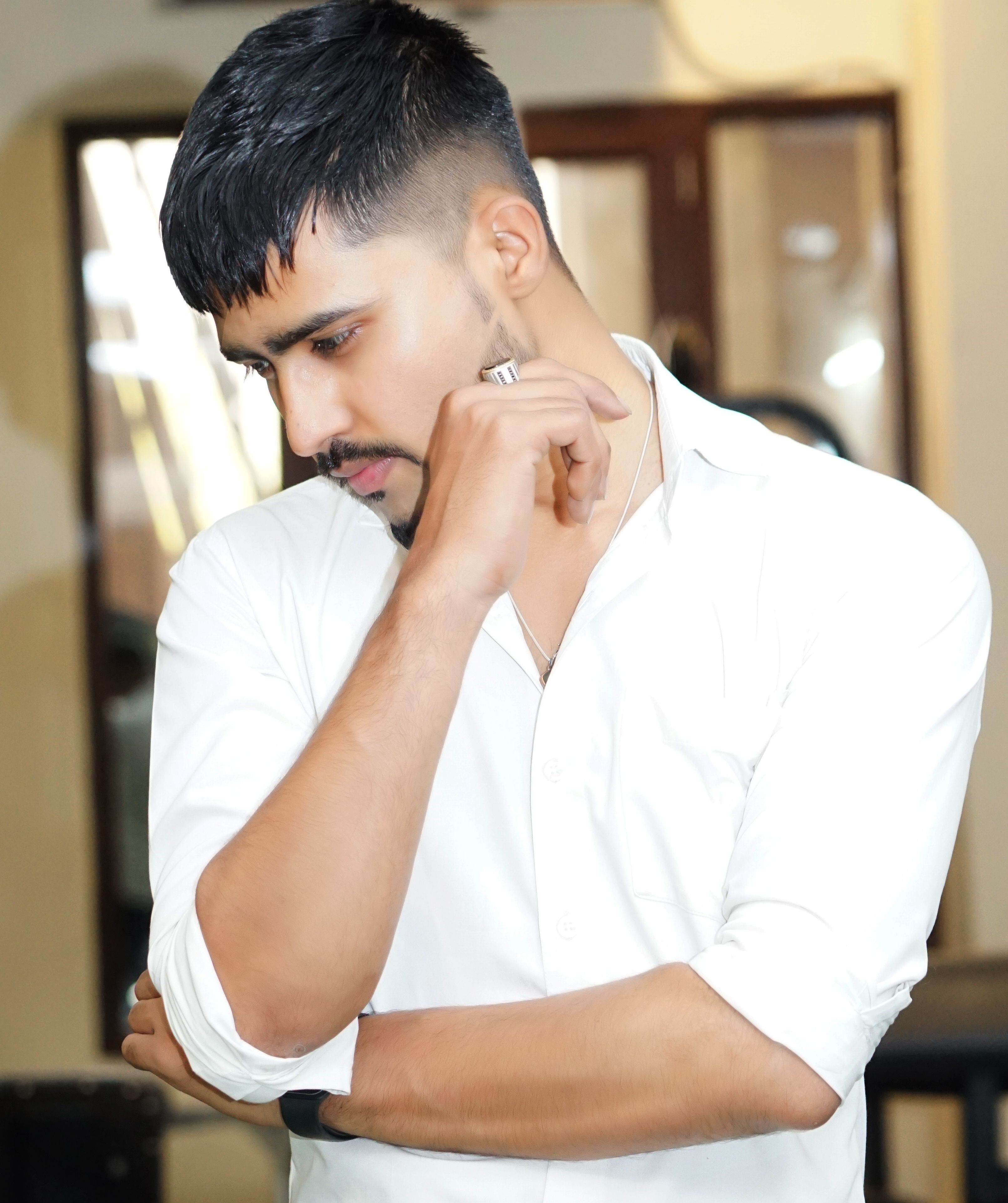 A young man in a white shirt appears thoughtful while standing in an indoor setting.