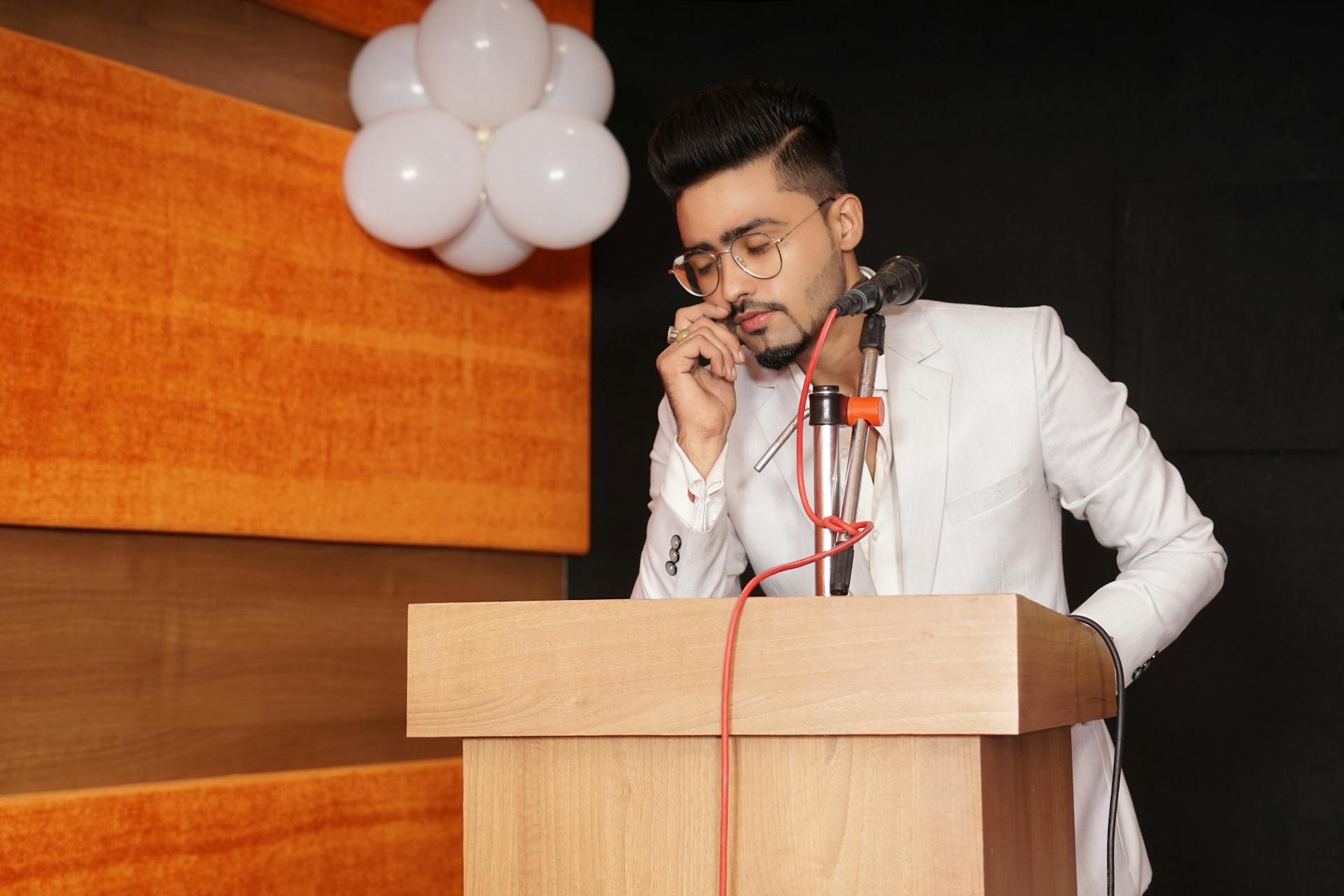 Young man in white suit speaks at a podium with balloons in background.