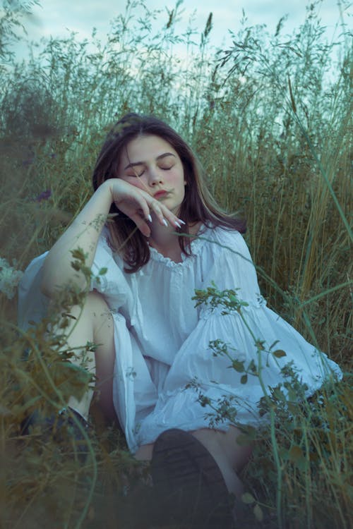 Woman With Closed Eyes in White Dress Sitting on Grass Field 