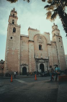 View of a historic cathedral in a sunlit town square with people and palm trees.