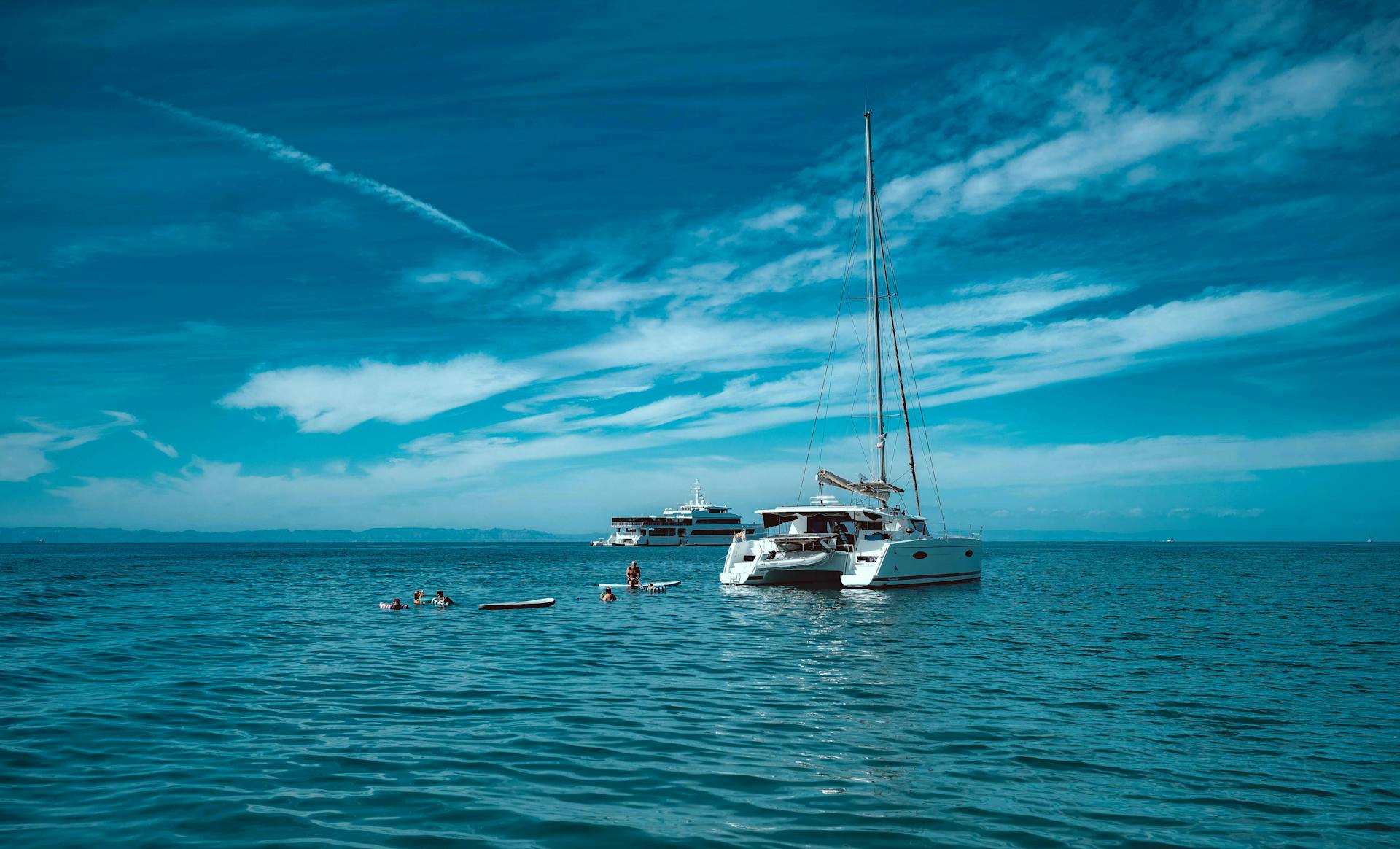 A catamaran sailing in open sea with people swimming nearby under a clear blue sky.