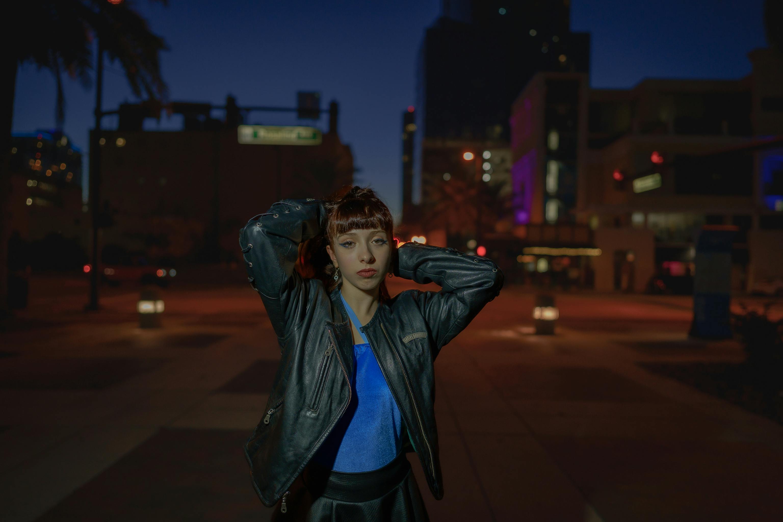 Free A young woman poses confidently on an illuminated city street at night, wearing a stylish leather jacket. Stock Photo