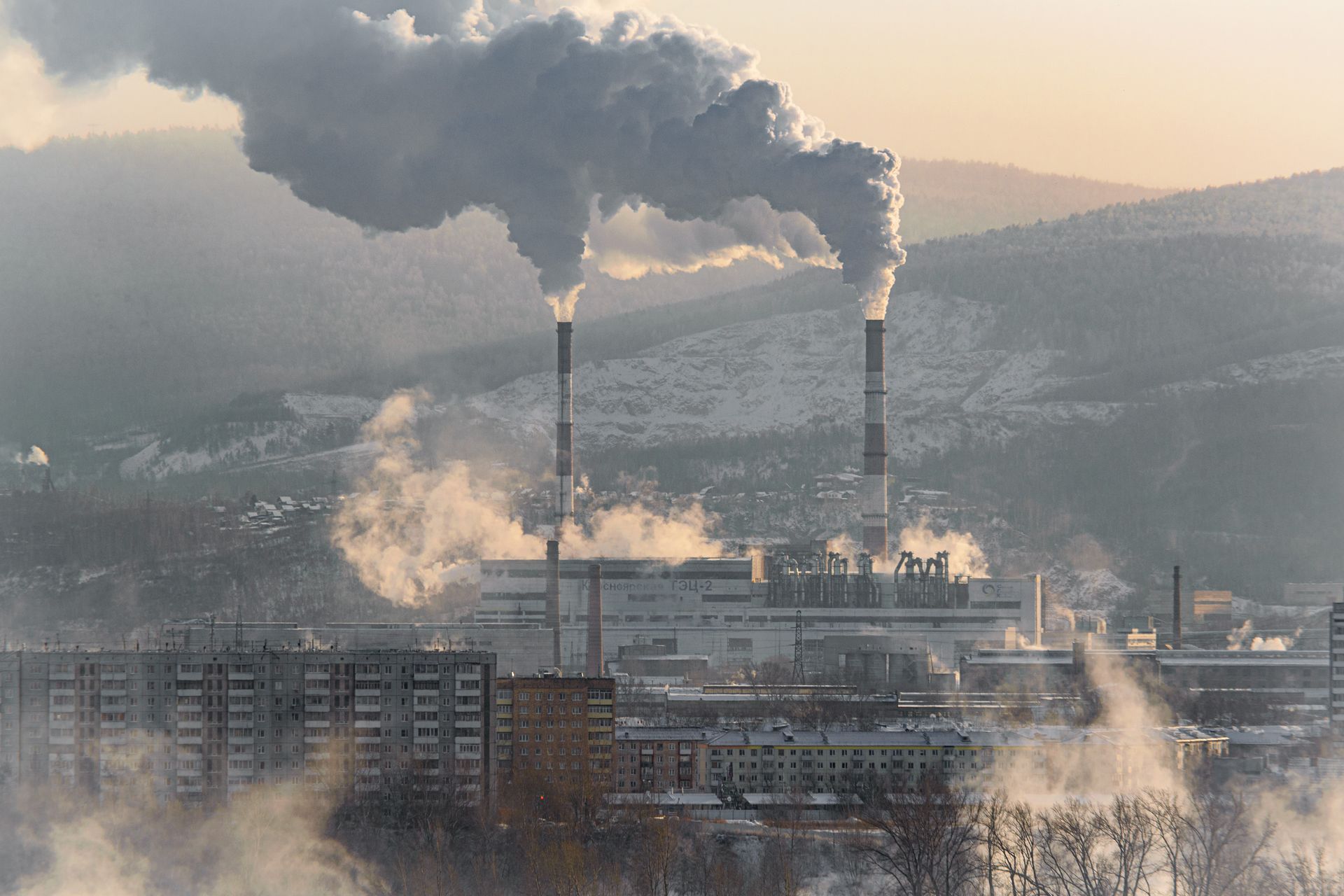 An industrial factory with chimneys emitting smoke, surrounded by snowy landscapes and buildings.