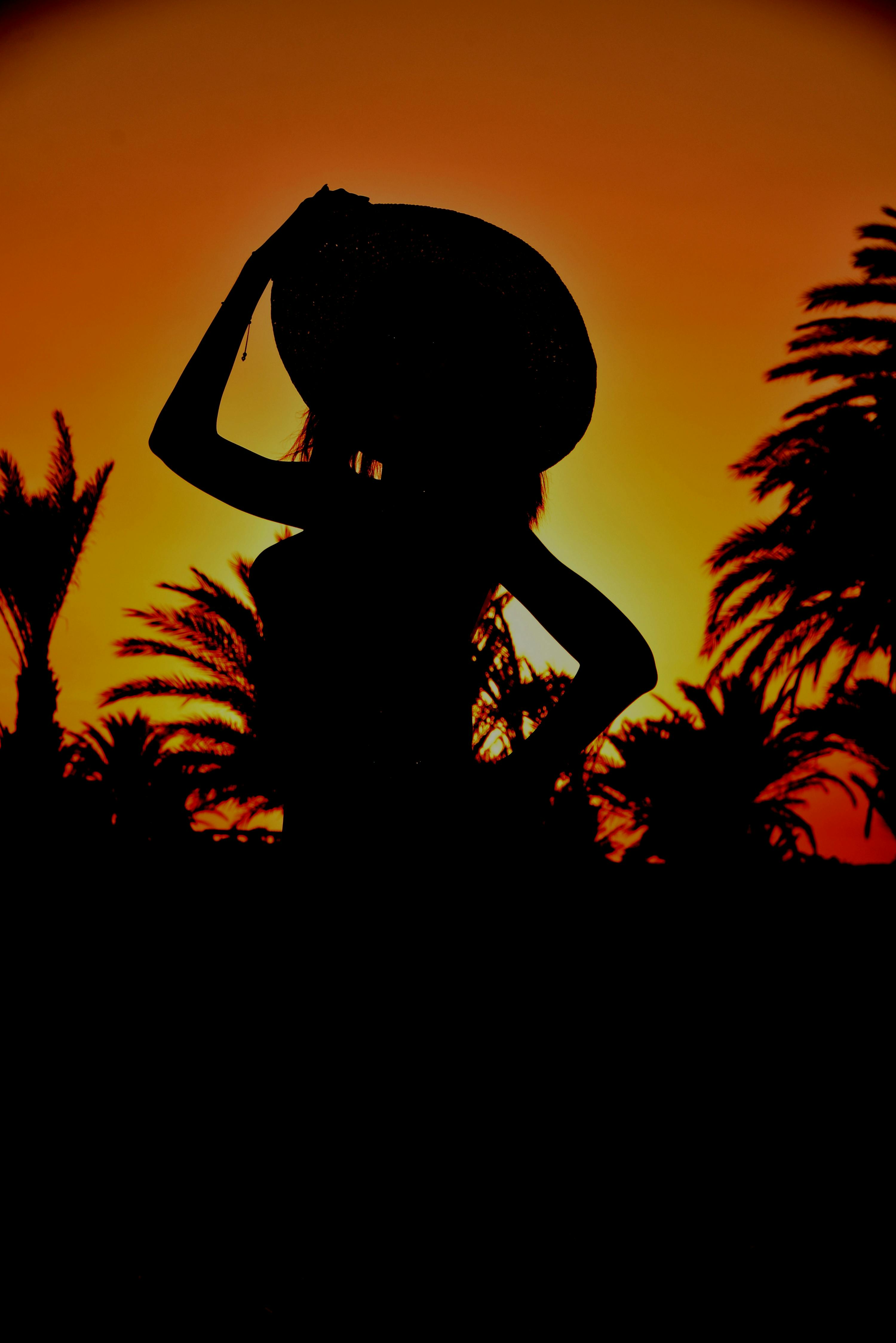 Stunning silhouette of a woman wearing a hat against a vibrant sunset sky with palm trees.