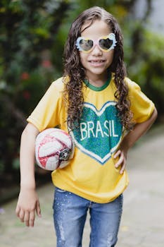 Cheerful child with curly hair wearing a Brasil shirt and sunglasses, holding a soccer ball outdoors.