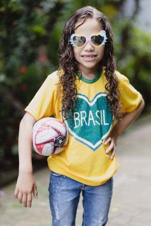 Free Cheerful child with curly hair wearing a Brasil shirt and sunglasses, holding a soccer ball outdoors. Stock Photo