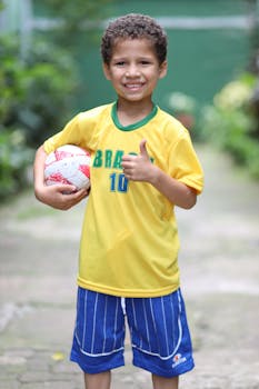Smiling young boy in Brazilian football jersey holding a soccer ball outdoors.