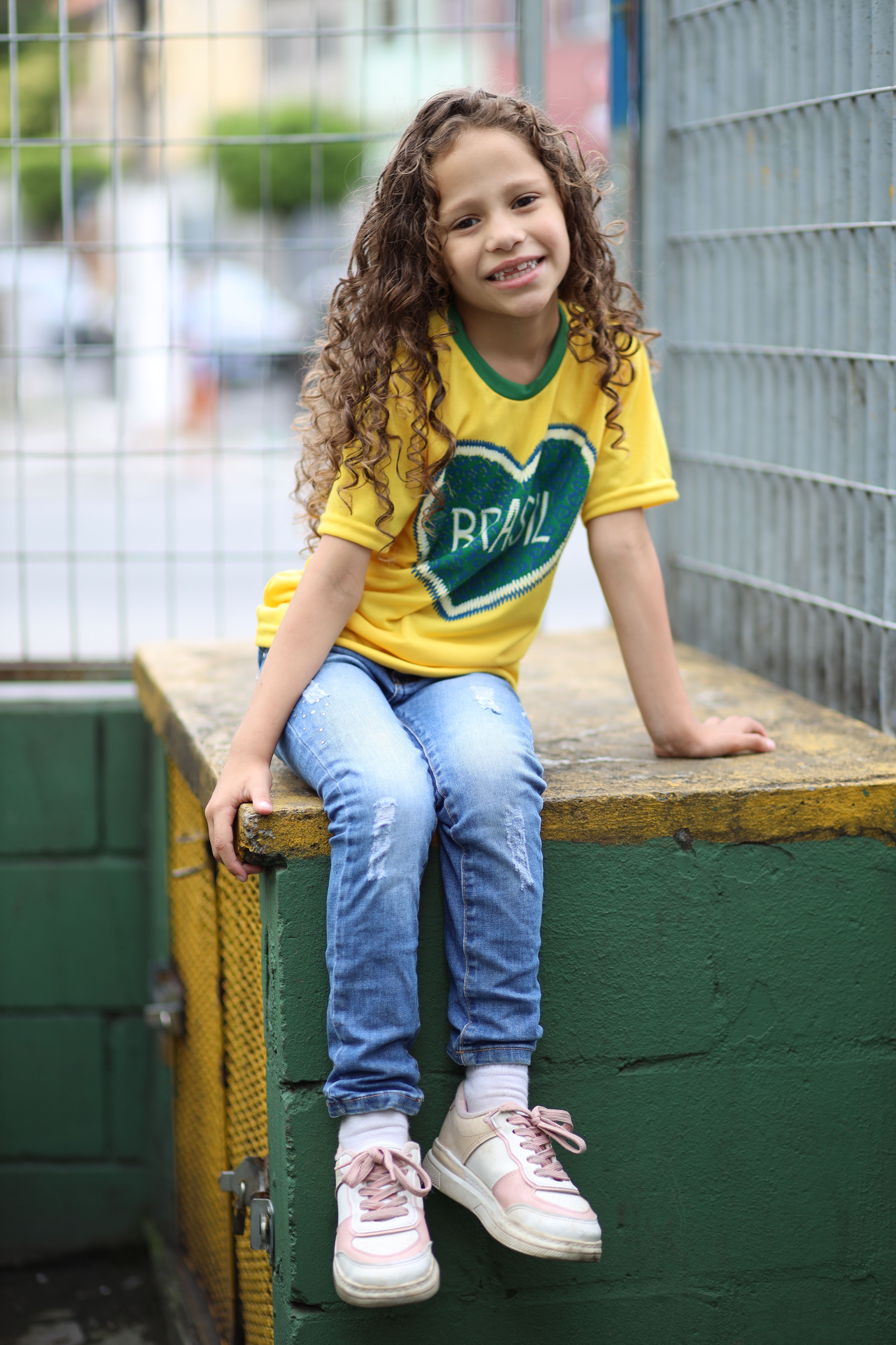 Happy girl with curly hair wearing a Brasil shirt, sitting outdoors on a sunny day.