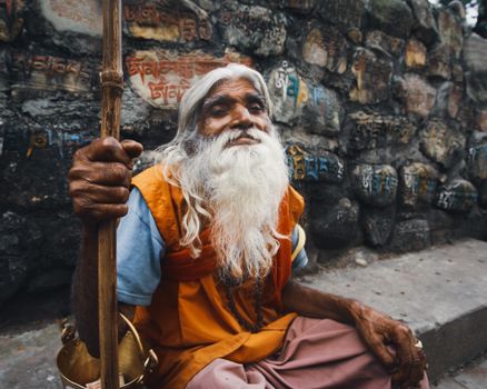 Portrait of an elderly monk with long beard wearing orange robes sitting near a temple shrine wall.