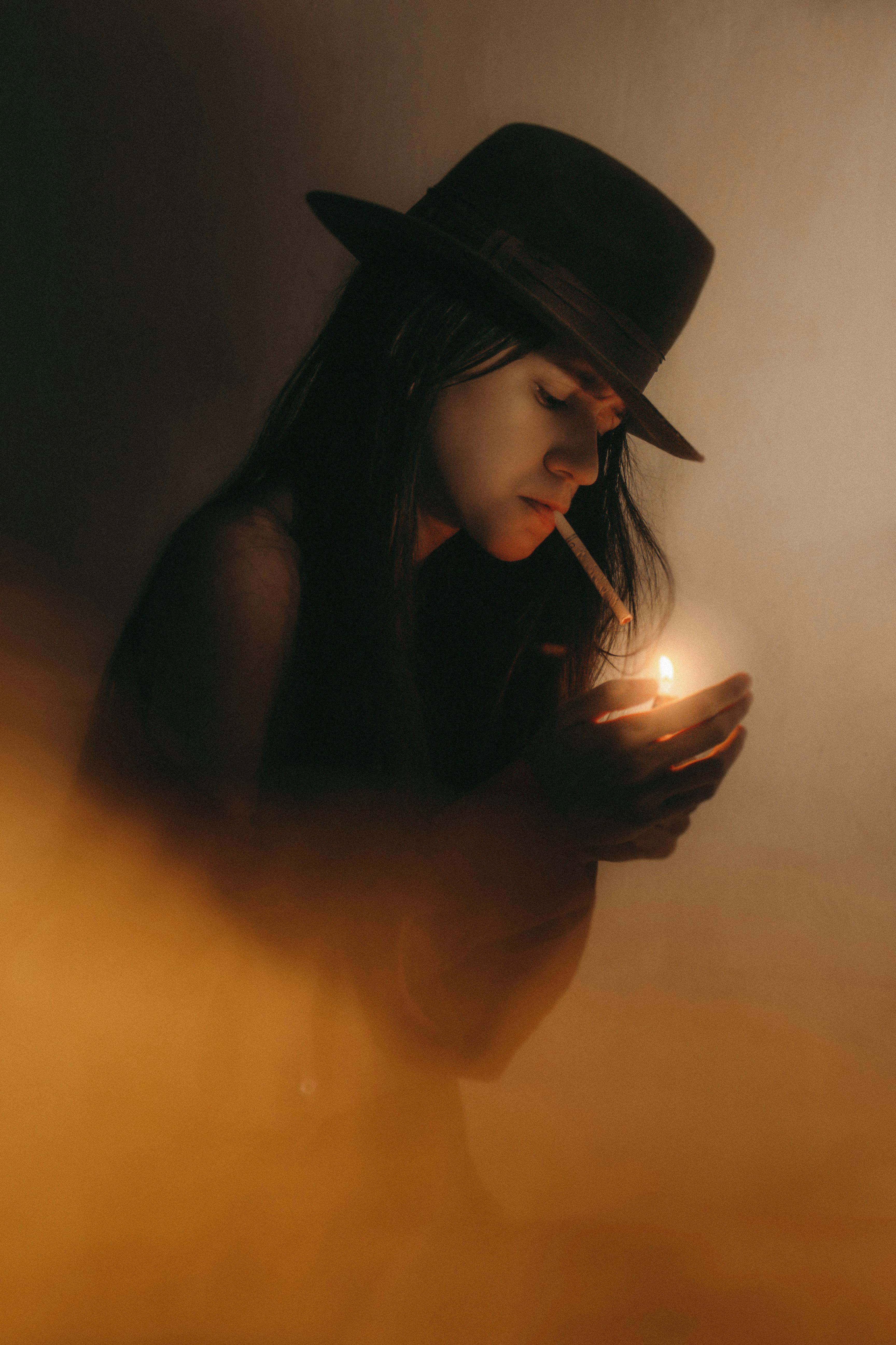 Artistic portrait of a young woman in a hat lighting a cigarette, captured in a moody atmosphere.