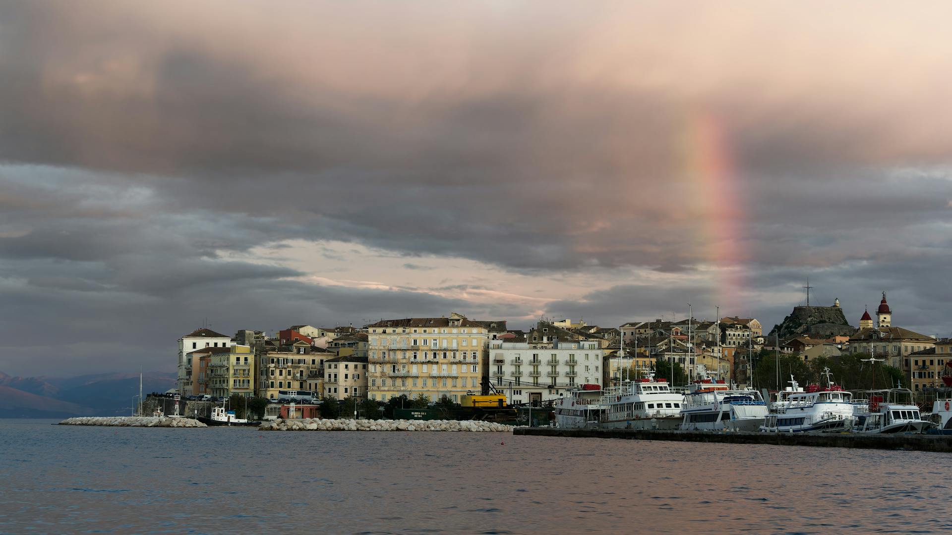 Dramatic Corfu cityscape featuring a vibrant rainbow over the harbor under overcast skies.
