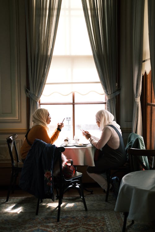 Women Sitting by a Table under a Window and Using Their Phones