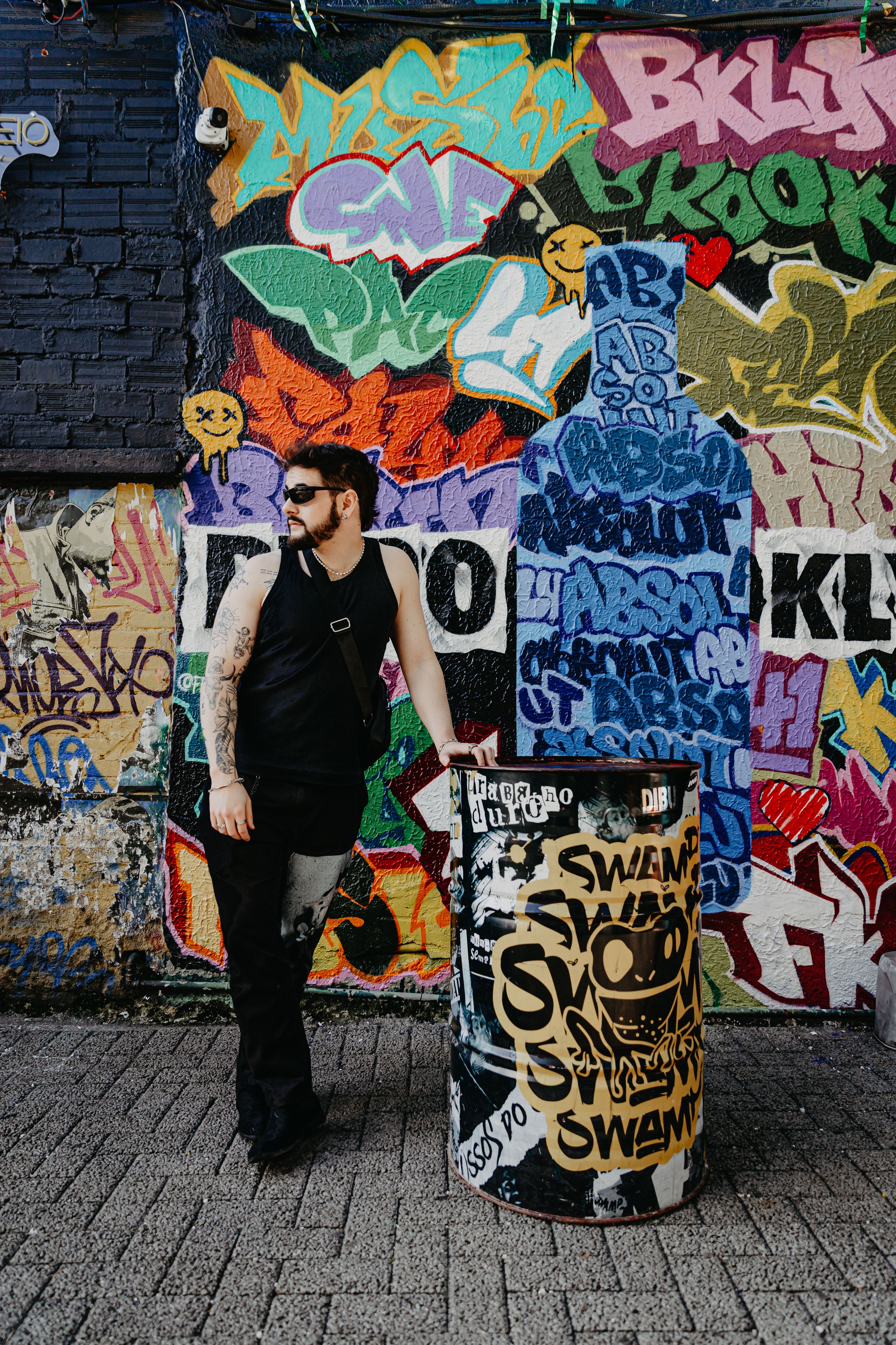Man in black outfit with sunglasses standing by graffiti wall, conveying urban street style.