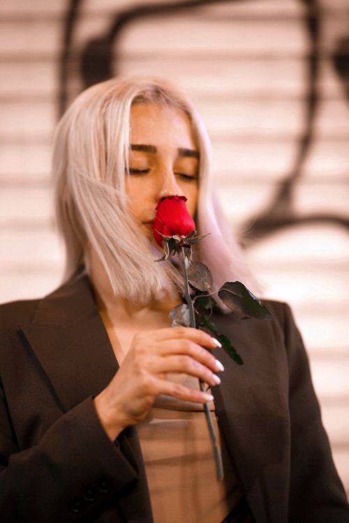 Free A woman in a black blazer intimately smelling a red rose with a serene expression. Stock Photo