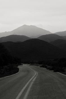 A dramatic black and white shot of a winding road leading to distant mountains under a moody sky.