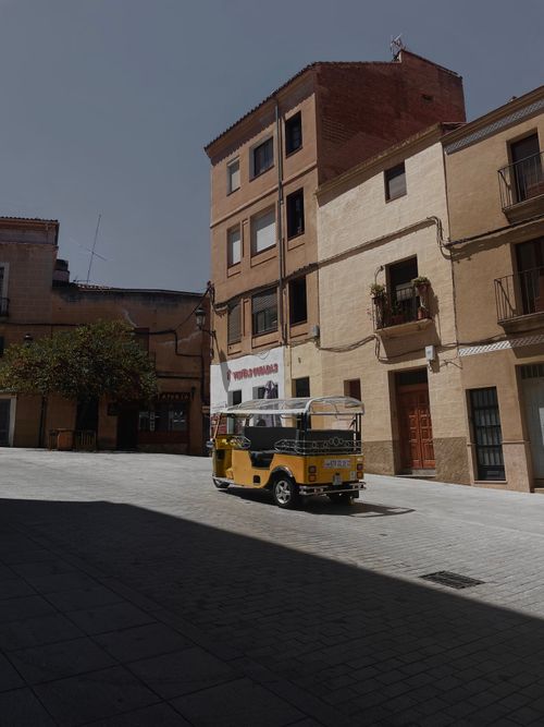 Free A yellow auto rickshaw parked on a quiet sunlit street surrounded by European-style buildings. Stock Photo