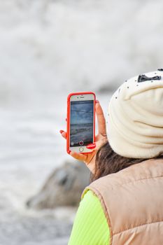 A woman in a beanie is taking photos of beach waves with her smartphone, showcasing a serene coastal scene in Georgia.
