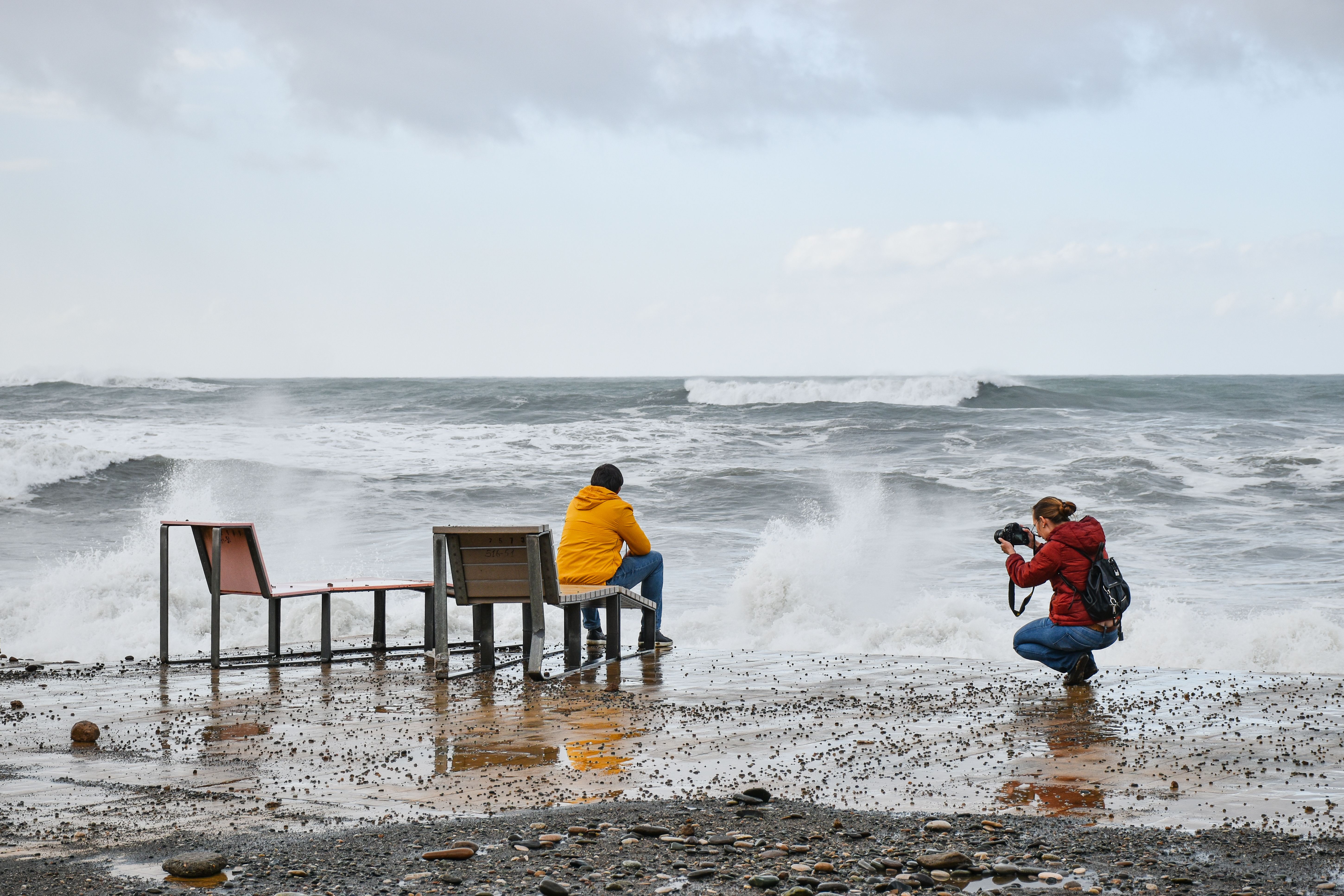 Free A photographer takes a picture of a person sitting by the sea amid crashing waves. Stock Photo