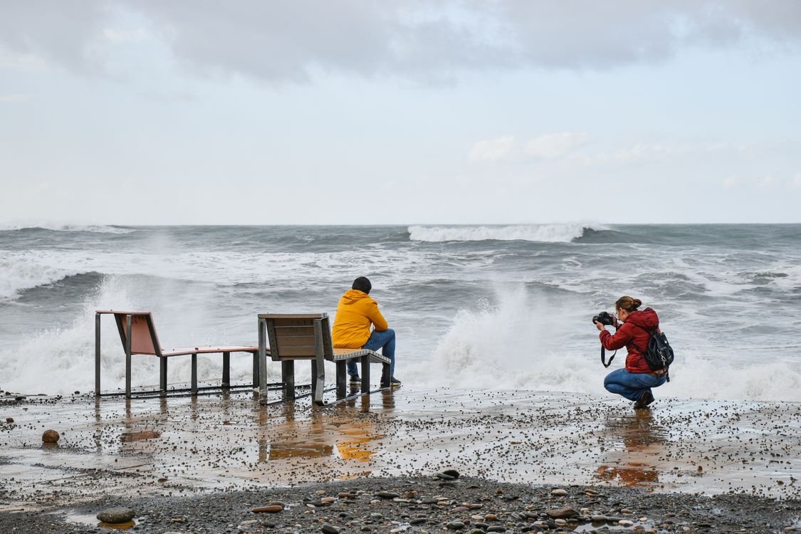 Free A photographer takes a picture of a person sitting by the sea amid crashing waves. Stock Photo