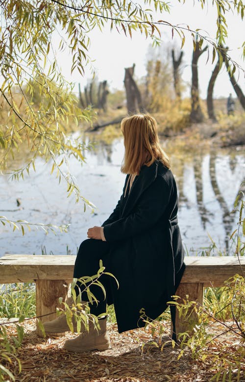 A Woman in a Black Coat Sitting on a Wooden Bench while Looking at a Lake