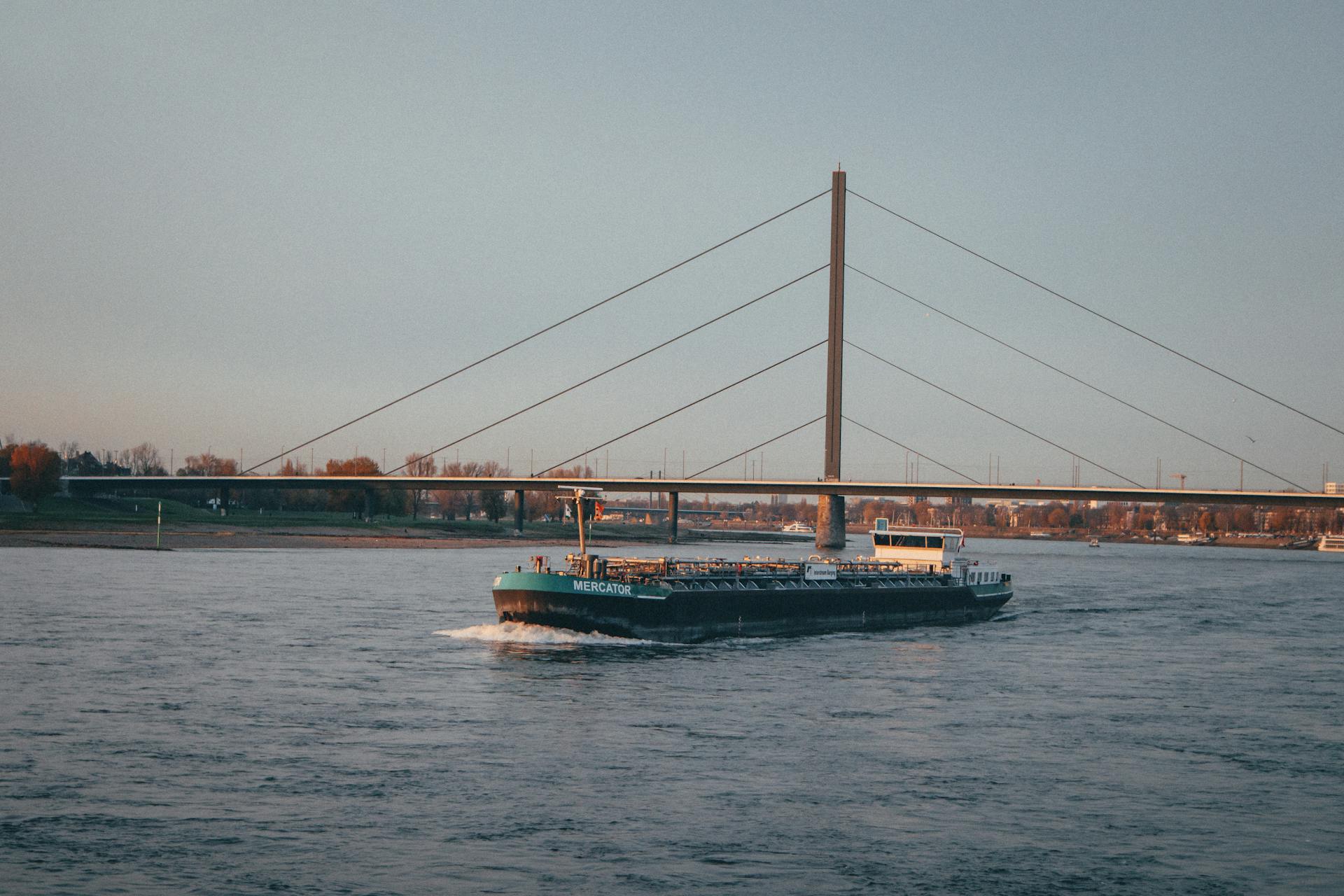 A cargo vessel passes under a modern suspension bridge on a tranquil river.