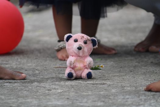 A dirty pink teddy bear sits on a concrete floor surrounded by children and a red balloon.