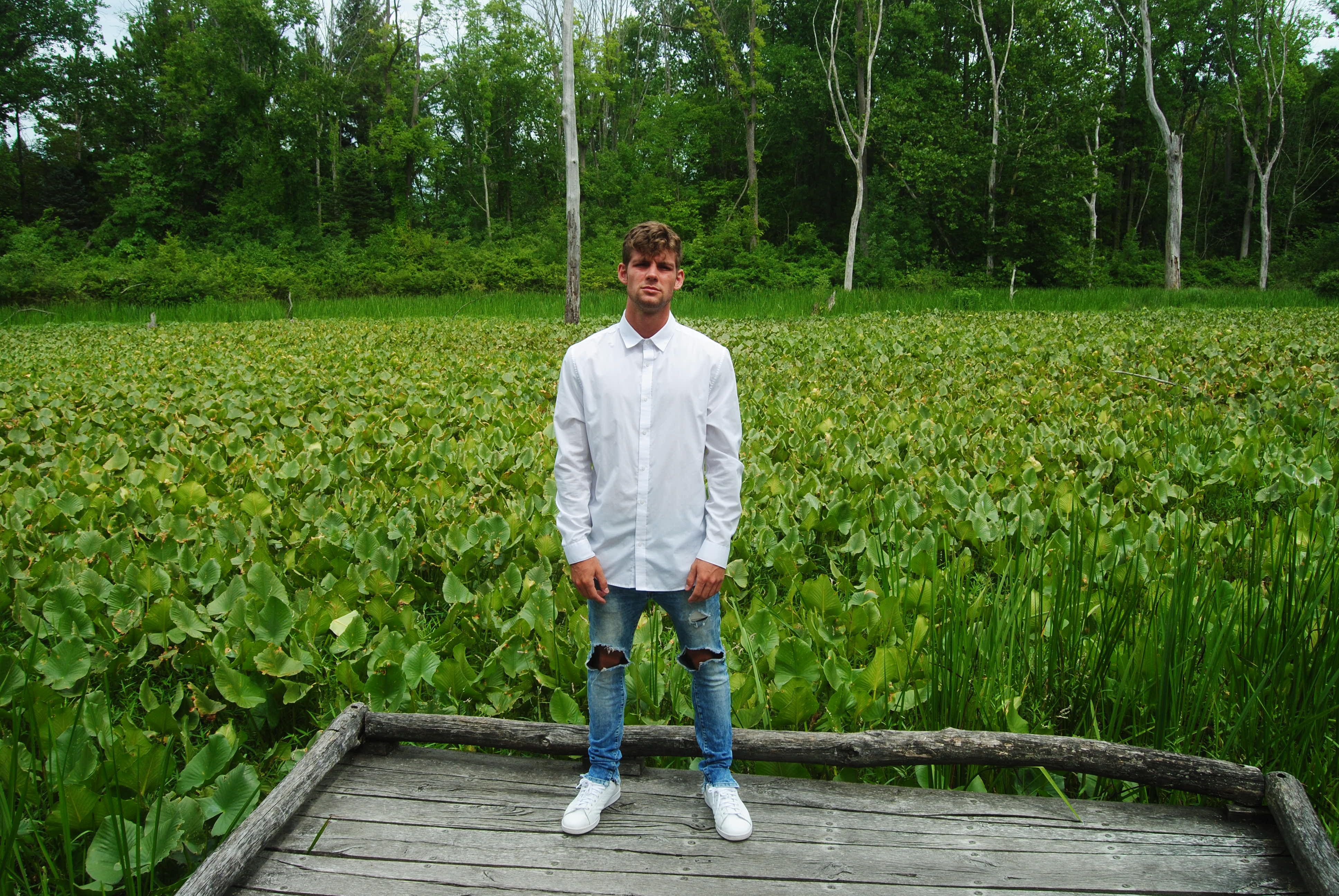 Young man in white shirt and jeans standing on a wooden pier in a lush marshland environment.