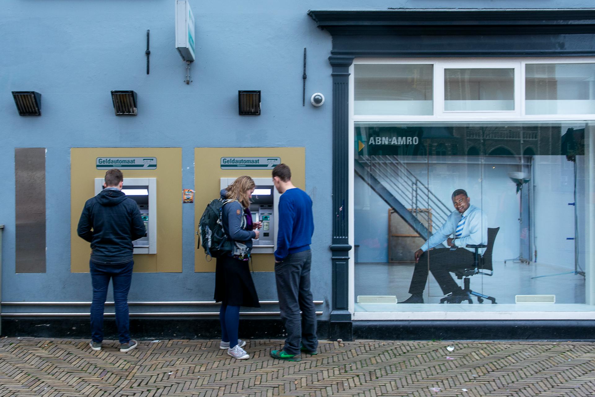 Two ATMs on city street with people using them and bank poster nearby.