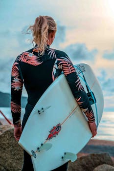 Female surfer holding a surfboard, wearing a floral wetsuit, enjoying a sunset at the beach.