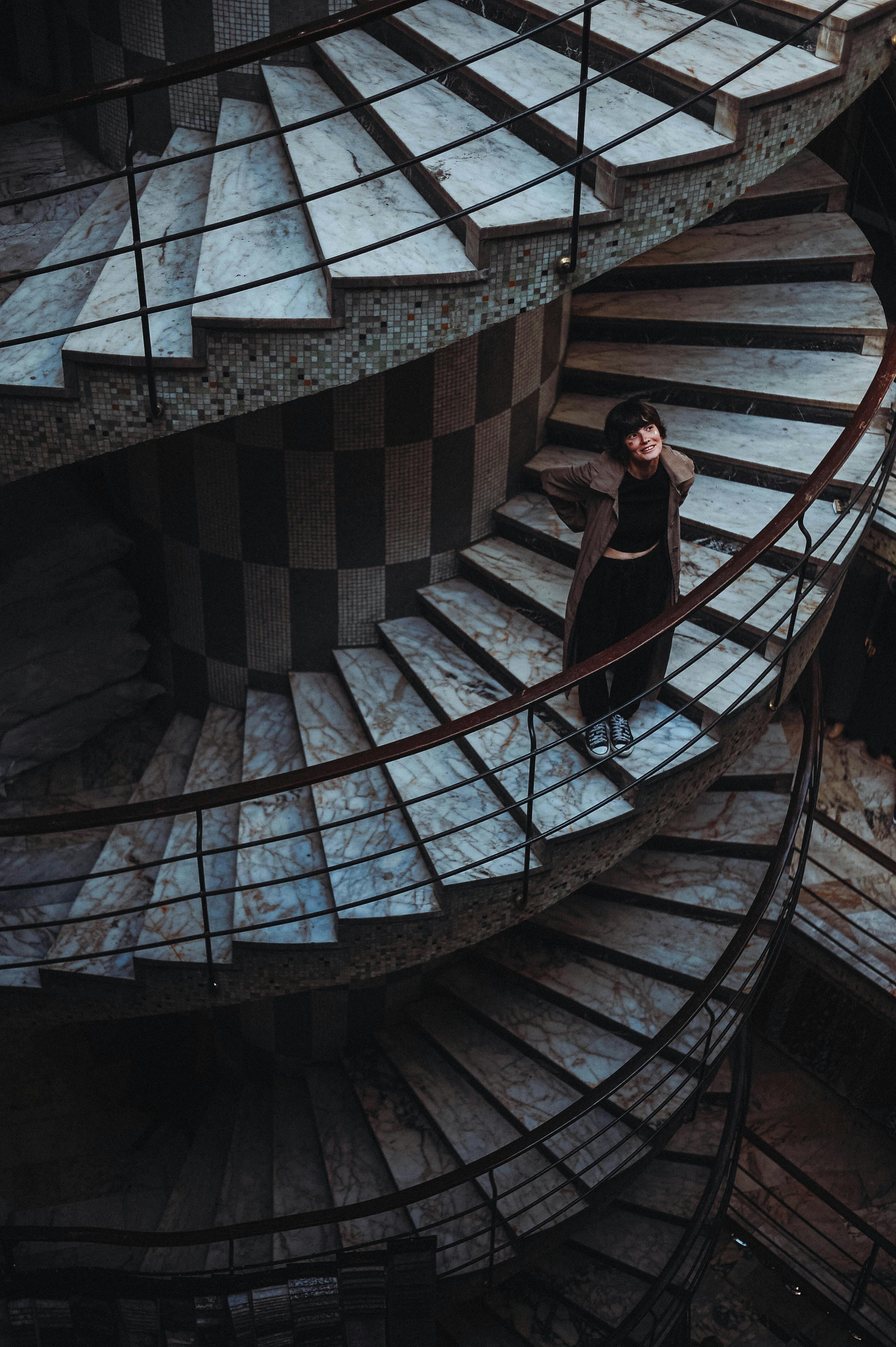 A woman in a coat stands indoors on a marble spiral staircase, captured from above.